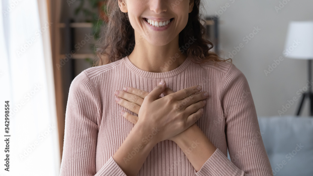 Close up focus on happy sincere female holding folded hands on chest. Emotional positive kind candid millennial woman feeling thankful indoors, showing gratitude sign, believe faith charity concept.