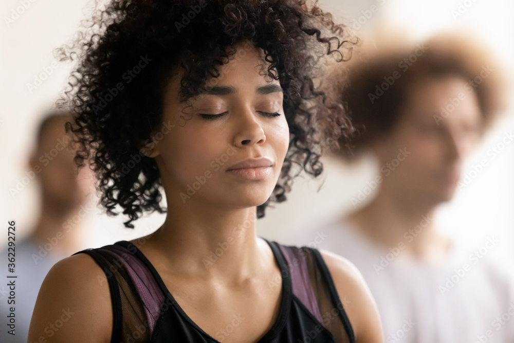 Head shot close up young peaceful attractive curly hair african american woman breathing fresh air, enjoying deep meditation with closed eyes, relaxing after yoga class workout in sport club.